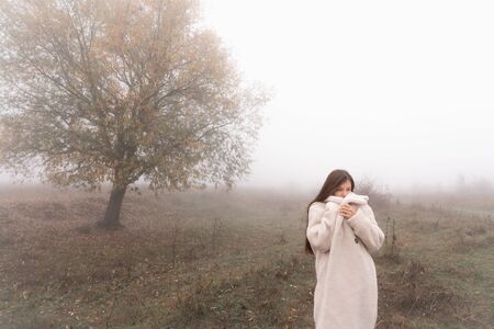 Attractive brunette girl wrapped in white coat stands among the meadow, big tree in fog on the backgroundの写真素材
