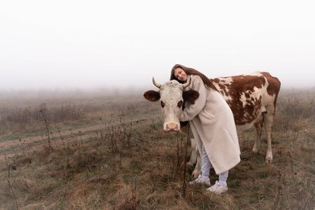 Beautiful brunette girl in white coat stands among the meadow in fog with red and white coe, hugging her and looking at the cameraの写真素材