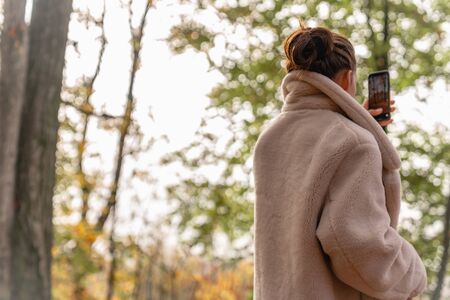 Attractive beautiful brunette girl in white coat walking among the colorful forest and making photo on her mobile phoneの写真素材