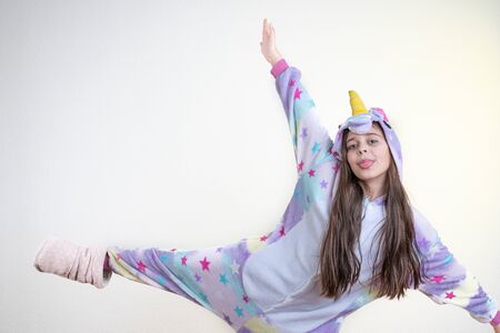 Playfull brunette child girl with loose hair dressed in unicorn pajamas stands on one leg with raised hands and other leg, shows tongue and looking at the camera, isolated over white backgroundの写真素材