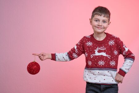 Attractive child boy dressed in red santa sweater, stands with raised hand and looking at the camera, holds red new years toy on her finger, isolated over pink background, Merry Christmas and Happy New Year conceptの写真素材