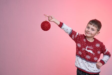Attractive child boy dressed in red santa sweater, stands with raised hand and looking at the camera, holds red new years toy on her finger, isolated over pink background, Merry Christmas and Happy New Year conceptの写真素材