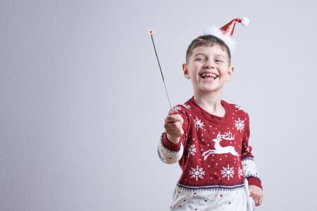 Blissful smiling child boy in red santa sweater and hat holds in his hand sparkler, isolated over grey background, Merry Christmas and Happy New Year conceptの写真素材