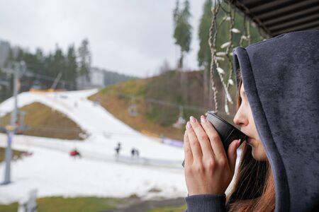 Close up portrait of the young female in hood drinking cofee or tee and looking away, mountains with snow on the backgroundの写真素材