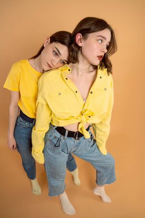 Upset, unhappy brunette girls in yellow shirts standing together isolated over orange backgroundの写真素材