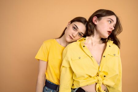 Two young brunette girls in yellow clothes over orange background, european woman leaned on her friend shoulder and looking at the cameraの写真素材