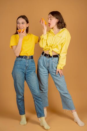 Two attractive brunette girls in casual clothes eating sliced orange while standing together against orange wallの写真素材