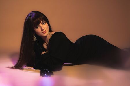 Fashion studio portrait of young beautiful female model lying in black sexy dress on orange...