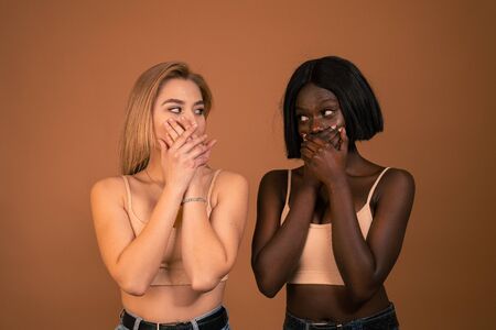 Waist up photo of two international girls in beige bra closing their mouths with their hands and looking at each otherの写真素材