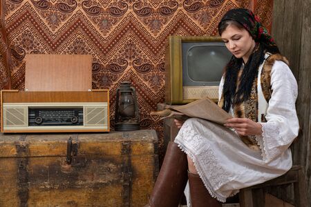 Serious woman in old national clothes sitting on the chair among vintage room and reading newspaper , retro tv, radio and gas lamp on the wooden chestの写真素材