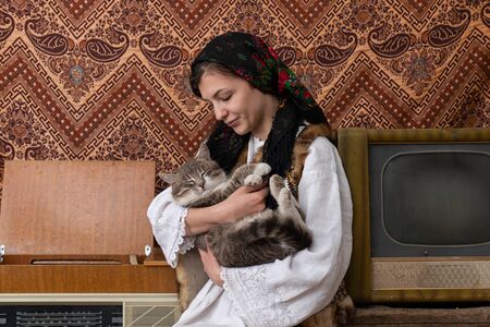 Young woman in national closes sitting among aged retro room and holding cat in her handsの写真素材