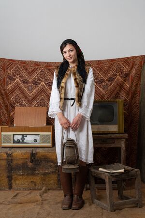 Cheerful young woman in national white dress and old leather shoes stands among vintage room, holds...