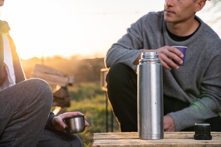 Wearing casual clothes man in drinking hot tea from a metal flusk while spending lunch break with girlfriend, sun lights on the backgroundの写真素材