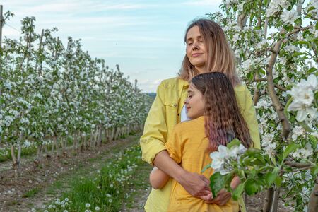 Curly brunette girl hugging her young mother, tender females with clothed eyes standing near blooming treesの写真素材