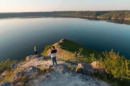 Young casual friends with phones standing on the top of the rock and looking at the wide river sunset in summerの写真素材