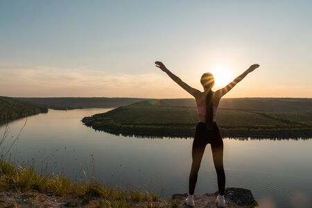 Silhouette of a sporty young woman standing on a ridge, enjoying the sunset over a river valley