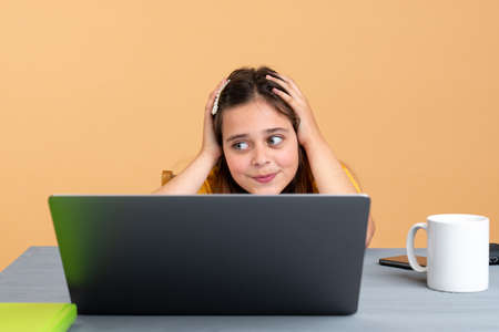 Exhausted schoolchild sitting at desk near laptop and copy book while doing schoolwork at home