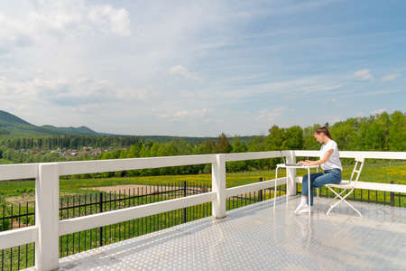 Concentrated stylish woman sitting on the white terrace balcony in a laptop apartment and working, staring intently at the screen. Beautiful girl freelancer working at a laptop in a country houseの写真素材