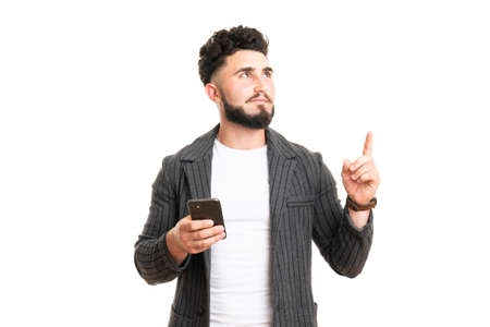 Portrait of an excited young man in casual using mobile phone while pointing finger up isolated over white backgroundの写真素材