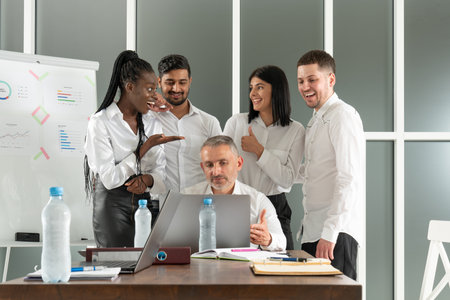 Male team leader listening to diverse colleagues questions, sitting together at table in office. Mixed race young teammates discussing project details at brainstorming meetingの写真素材