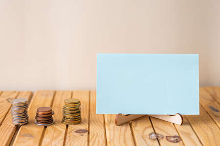 Empty Piece Of Paper On Holder Beside Stockpile Of Coins Over Desk. Blank Note Standing With Stacks Of Coin Placed On Wooden Table.の写真素材