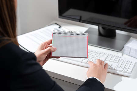 Businesswoman Holding Important Message Wtitten On Notebook On Desk With Computer, Clipboard And Notes. Crutial Information Presented On Notepad On Table With Keyboard And Pc.の写真素材