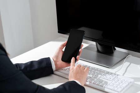 Businesswoman Holding Mobile Phone With Important Messages And Pointing On It Sitting On Desk. Woman Having Cellphone With New Ideas In Hand On Table With Computer.の写真素材