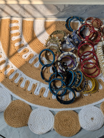 A stunning overhead shot of a colorful assortment of ethnic bangles and accessories arranged on a handmade jute rug with decorative circular mats. The bangles come in various colors including blue, red, gold, black, and adorned with intricate patterns and beads. The textured weave of the rug is clearly visible, featuring white and tan tones with a woven border. Sunlight casts shadows through window blinds creating an artistic pattern on the surface. This image captures vibrant ethnic jewelry for commercial use, ideal for showcasing traditional crafts in retail or online marketplaces.