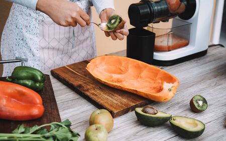 Young woman preparing organic juice with cold pressed extractor machine - Girl making smoothie with vegetables and fruit - Healthy nutrition during isolation quarantine - Focus on hand holding kiwiの写真素材
