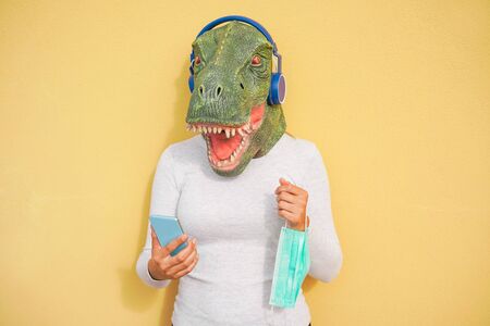Crazy woman wearing t-rex and holding protection mask while listening music with phone app - Quarantine isolation lifestyle during coronavirus time - Absurd and funny trend concept - Focus on faceの写真素材
