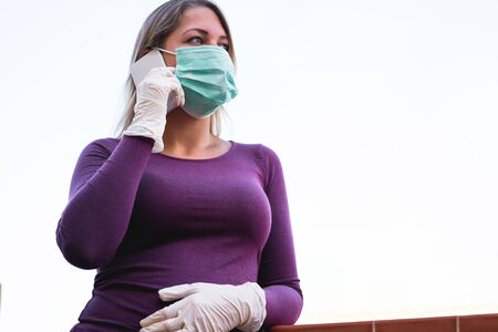Young woman talking with mobile phone outdoor wearing face mask and gloves for Covid-19 virus prevention - Girl speaking with smartphone outside during quarantine isolation - Focus on left hand gloveの写真素材