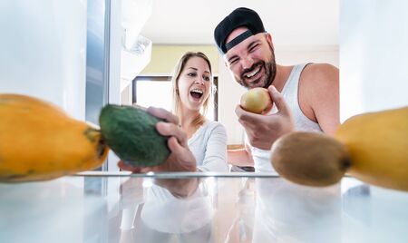 Young couple open the fridge at home while choosing organic fruits for fresh smoothie - Happy people having fun cooking inside kitchen - Helathy food and relationship concept - Focus on blond girlの写真素材