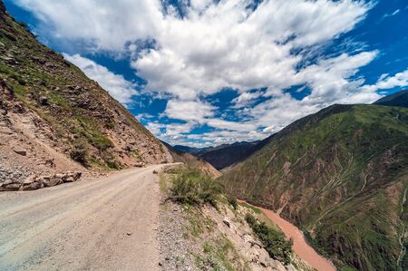 Mountain dirt road with no guardrail or protection overlooking a very deep valley with a river under a blue sky with white clouds in the Tibetan Himalayasの写真素材