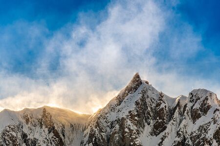 Wind blows snow from the mountain tops creating a white cloud illuminated by the sun against the...