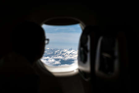 Silhouette of person with glasses looking out the window of an airplane overflying the Alpsの写真素材