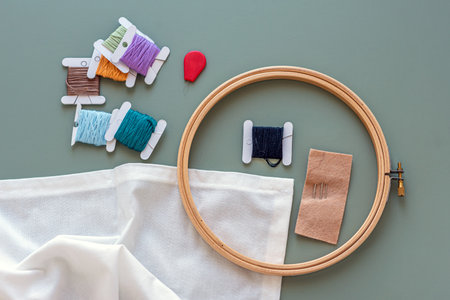 Embroidery tools on green background with wooden embroidery hoop, needles, needle threader and...