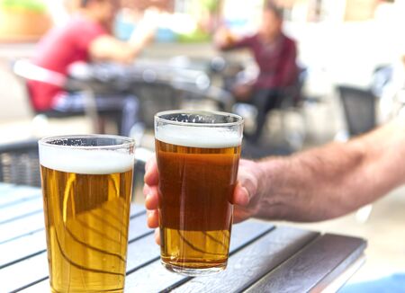 male hand  holding beer glass at table out doors out of focus drinkers in backgroundの写真素材
