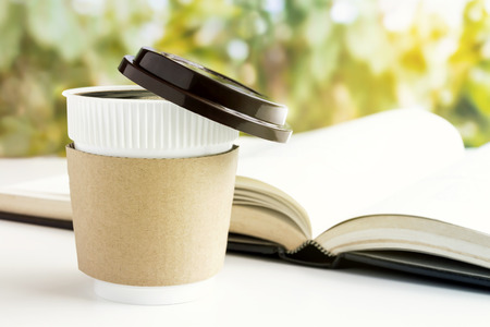 Paper coffee cup or disposable cup and textbook on the white table on green natural background with sunshine morning. With copy space for your text and logo.の写真素材
