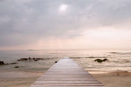 Wood bridge on the sea which has walk way for travel tourism and beautiful sky with sunshine in the morning. Sea waves slowly a the splashing on the sand and rocks with moss background.の写真素材