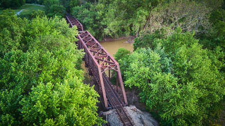 Aerial view above large metal bridge for train tracks over river surrounded by green treesの写真素材