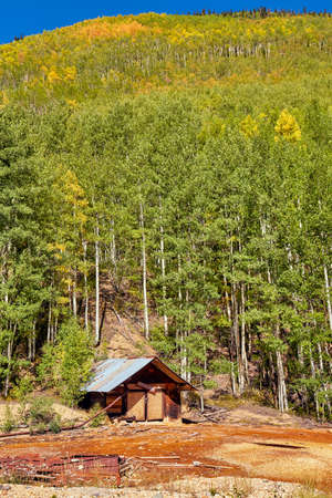 Small abandoned shack at base of large hill covered in green and yellow fall aspen treesの写真素材