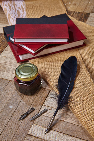 Black feather quill with additional nibs lays next to a closed ink pot and three red and black journalsの写真素材