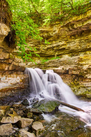 Cliff with waterfall creating gorge as water splashes against boulders and logの写真素材