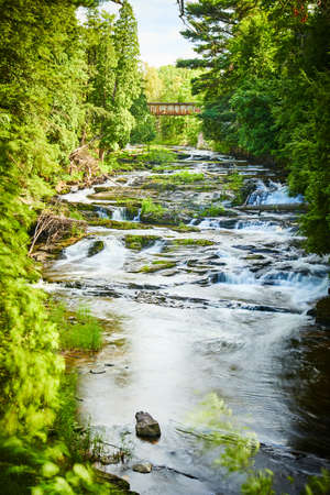 View through trees of waterfalls cascading down river with bridge in distanceの写真素材