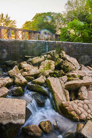 Pile of stone with small waterfall from dam and railroad bridge in background during sunset or sunriseの写真素材
