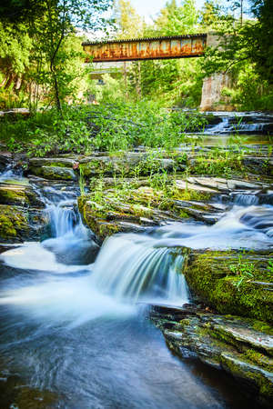 Old rusted bridge above beautiful cascading waterfalls over mossy rocksの写真素材