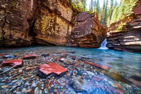 Red and blue river rocks in shallow river at bottom of gorge with waterfall and steep cliffsの写真素材