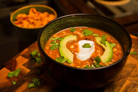 Delicious chicken enchilada soup with cilantro sour cream and avocado in a black bowl on a wooden cutting boardの写真素材