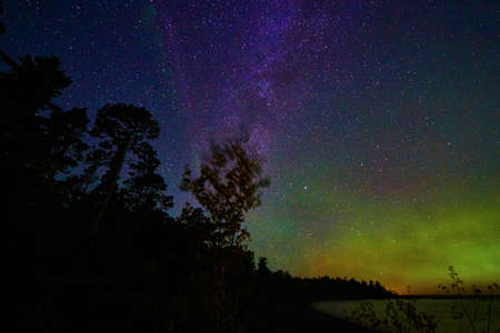 Deep blue and violet night sky with water and green and orange glow of aurora lights with tree silhouettesの写真素材