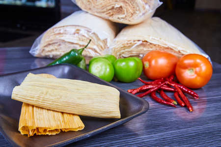 Tamales on tray with mexican ingredients of corn husks, tomatoes, and peppers in backgroundの写真素材